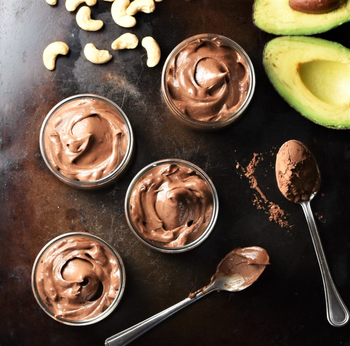 4 round chocolate mousse pots, 2 spoons with cocoa powder, cashew nuts and halved avocado.