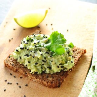 Avocado mixture with cilantro leaf on dark bread with lime wedge in background on light wooden cutting board.
