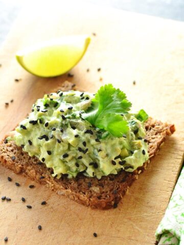 Avocado mixture with cilantro leaf on dark bread with lime wedge in background on light wooden cutting board.