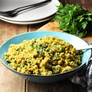 Close-up view of curried quinoa with peas in blue bowl with spoon, 2 white plates with forks and fresh herbs in background.