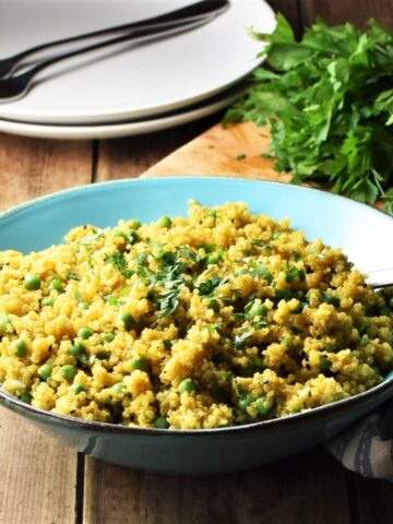 Close-up view of curried quinoa with peas in blue bowl with spoon, 2 white plates with forks and fresh herbs in background.