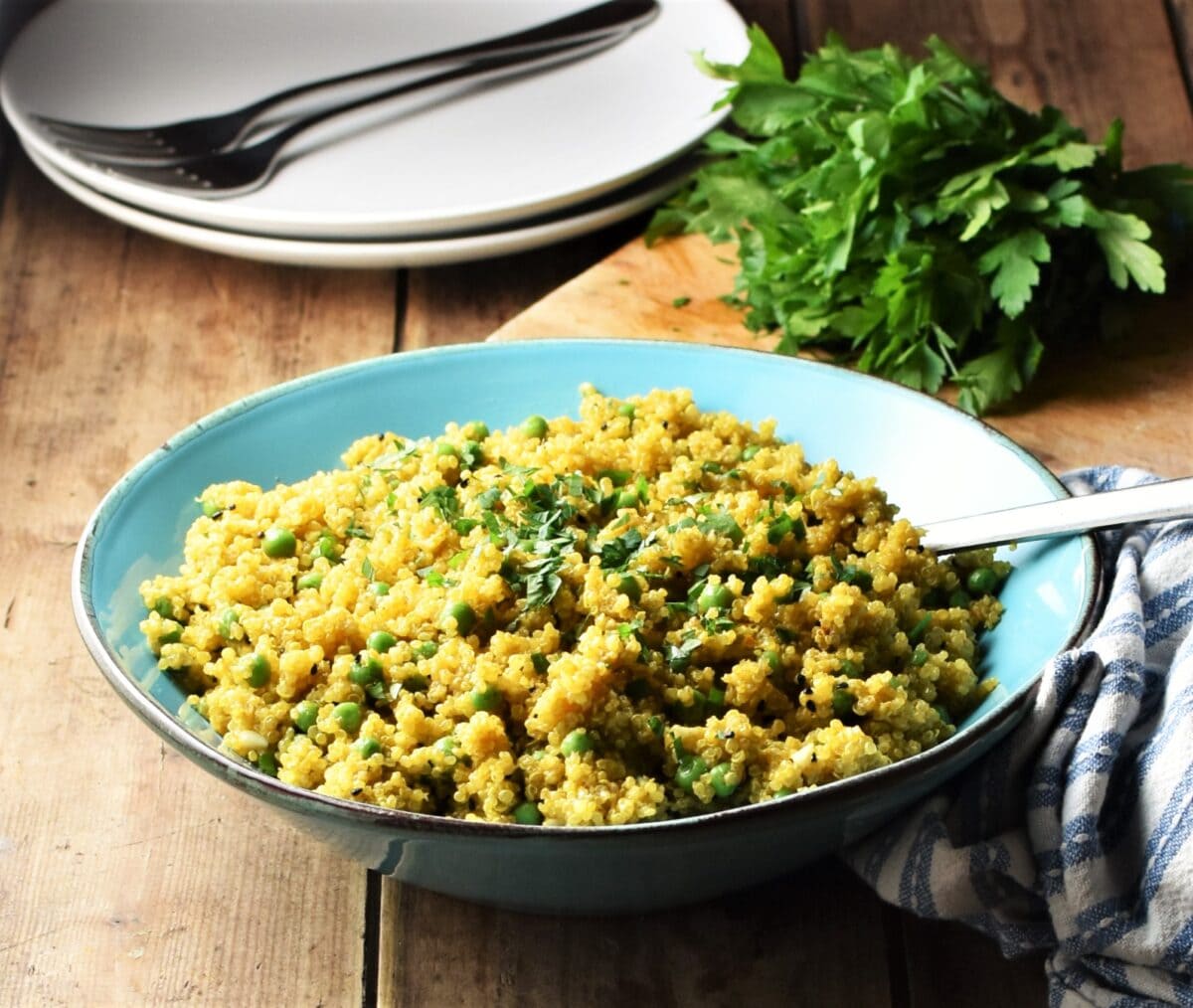 Close-up view of curried quinoa with peas in blue bowl with spoon, 2 white plates with forks and fresh herbs in background.