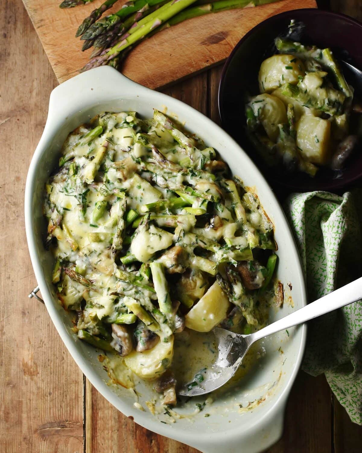Asparagus, potatoes and mushrooms in creamy sauce with spoon in green oval dish, with green cloth to the right, potato mixture in dish and asparagus tips in background.