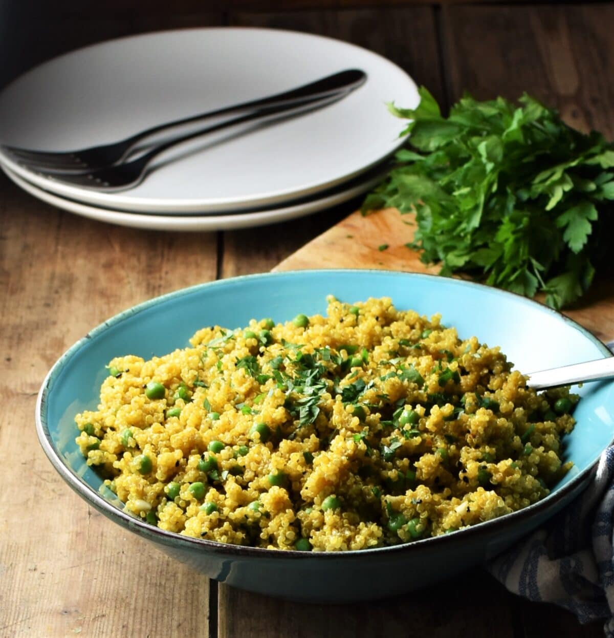 Curry quinoa with peas in blue bowl with spoon, 2 white plates with forks and fresh herbs in background.