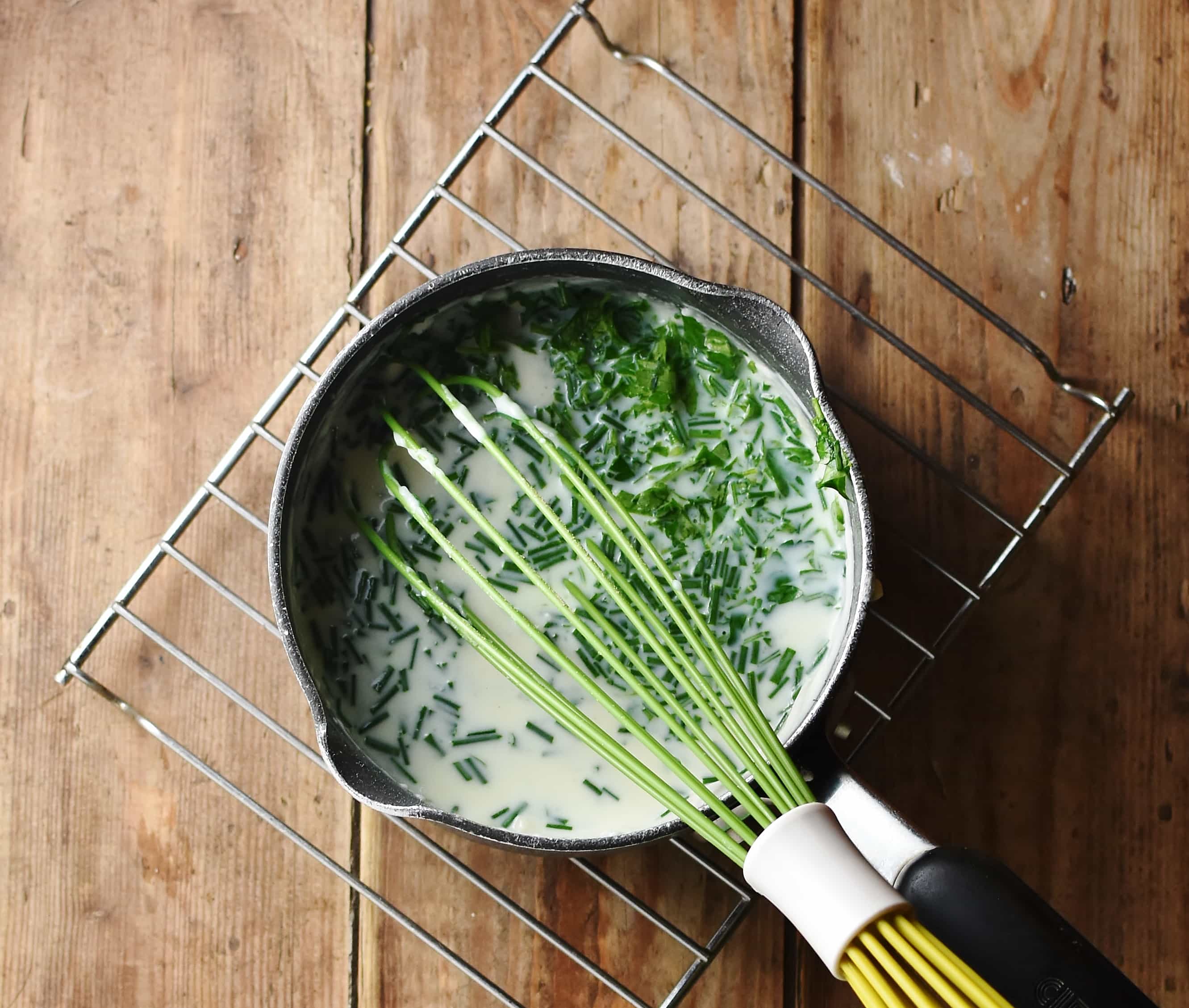 Sauce with herbs and green whisk in saucepan.