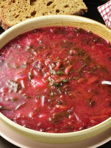 Side view of beet greens soup in beige bowl with spoon and bread in background.