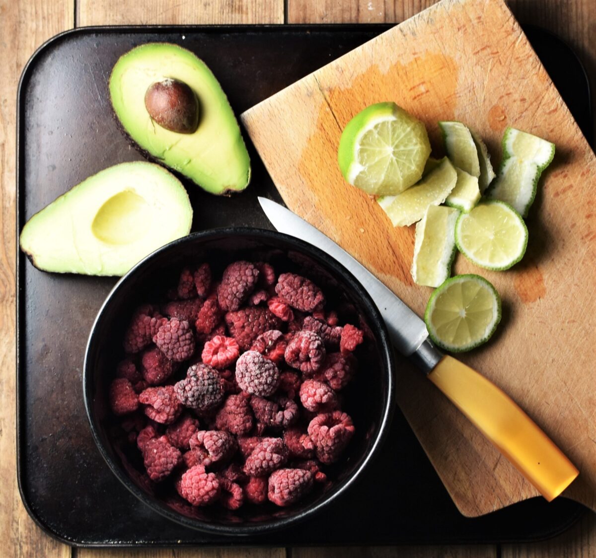 Halved avocado, peeled lime on top of chopping board with yellow knife and frozen raspberries in purple bowl.