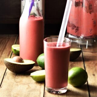 Side view of raspberry avocado smoothie in glass with straw with avocado, limes, smoothie in bottle and blender in background.