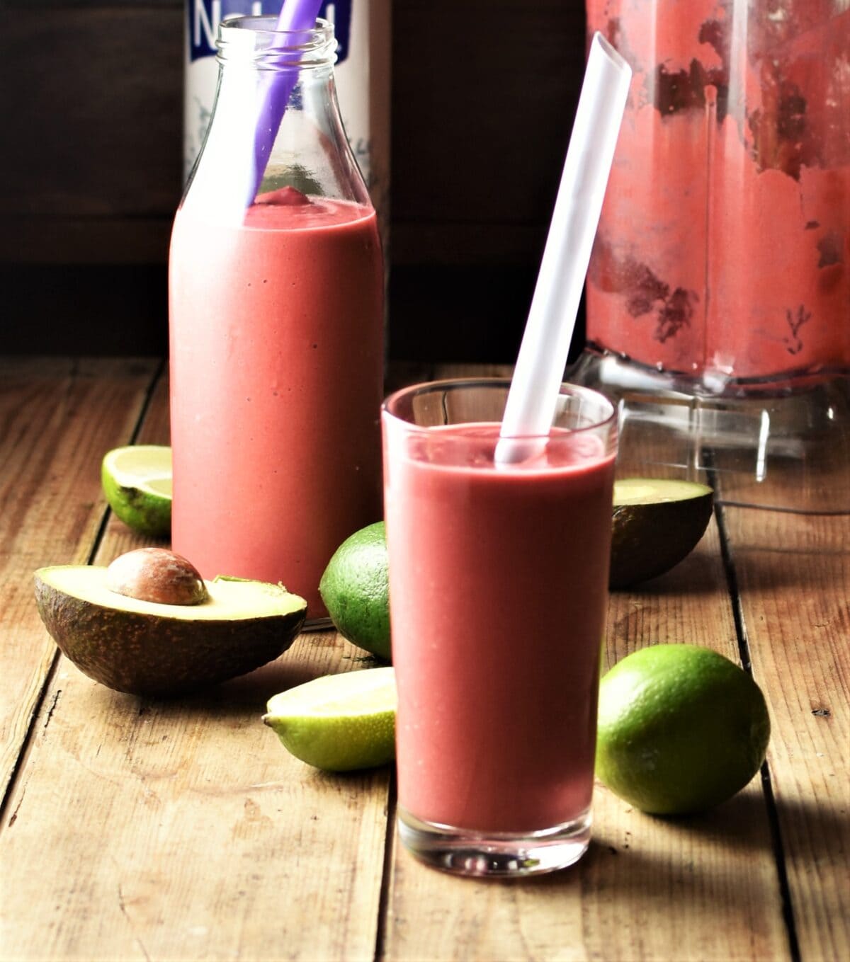 Side view of raspberry avocado smoothie in glass with straw with avocado, limes, smoothie in bottle and blender in background.