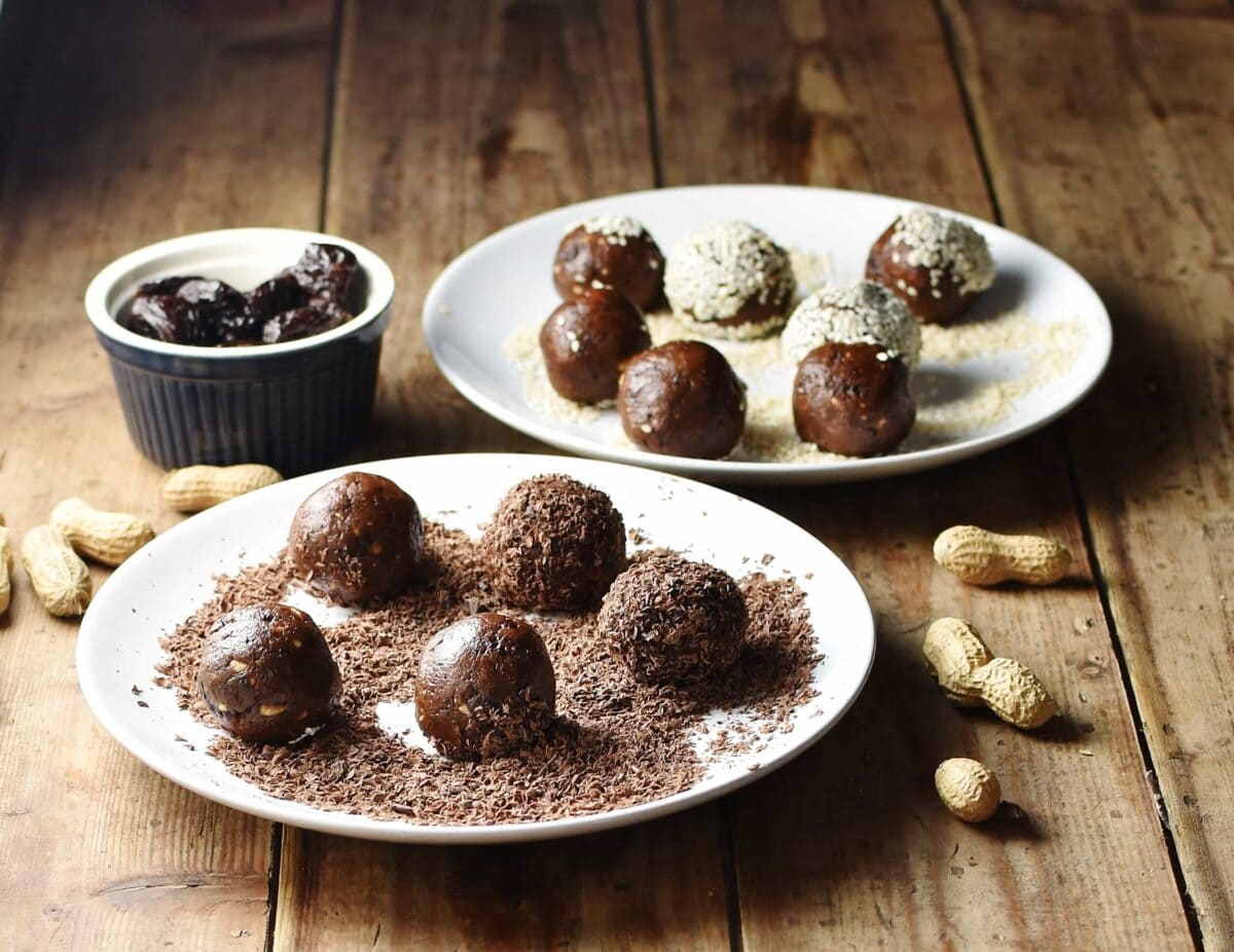 Side view of energy balls on 2 white plates coated in chocolate and seeds, with peanuts and prunes in blue container in background.