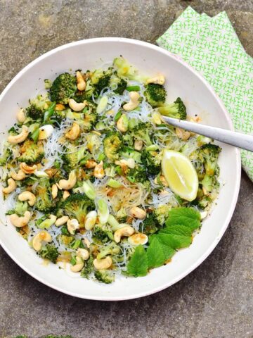 Top down view of bean thread noodles broccoli salad with cashews, lemon wedge, mint leaves and fork in white bowl with green cloth to right.