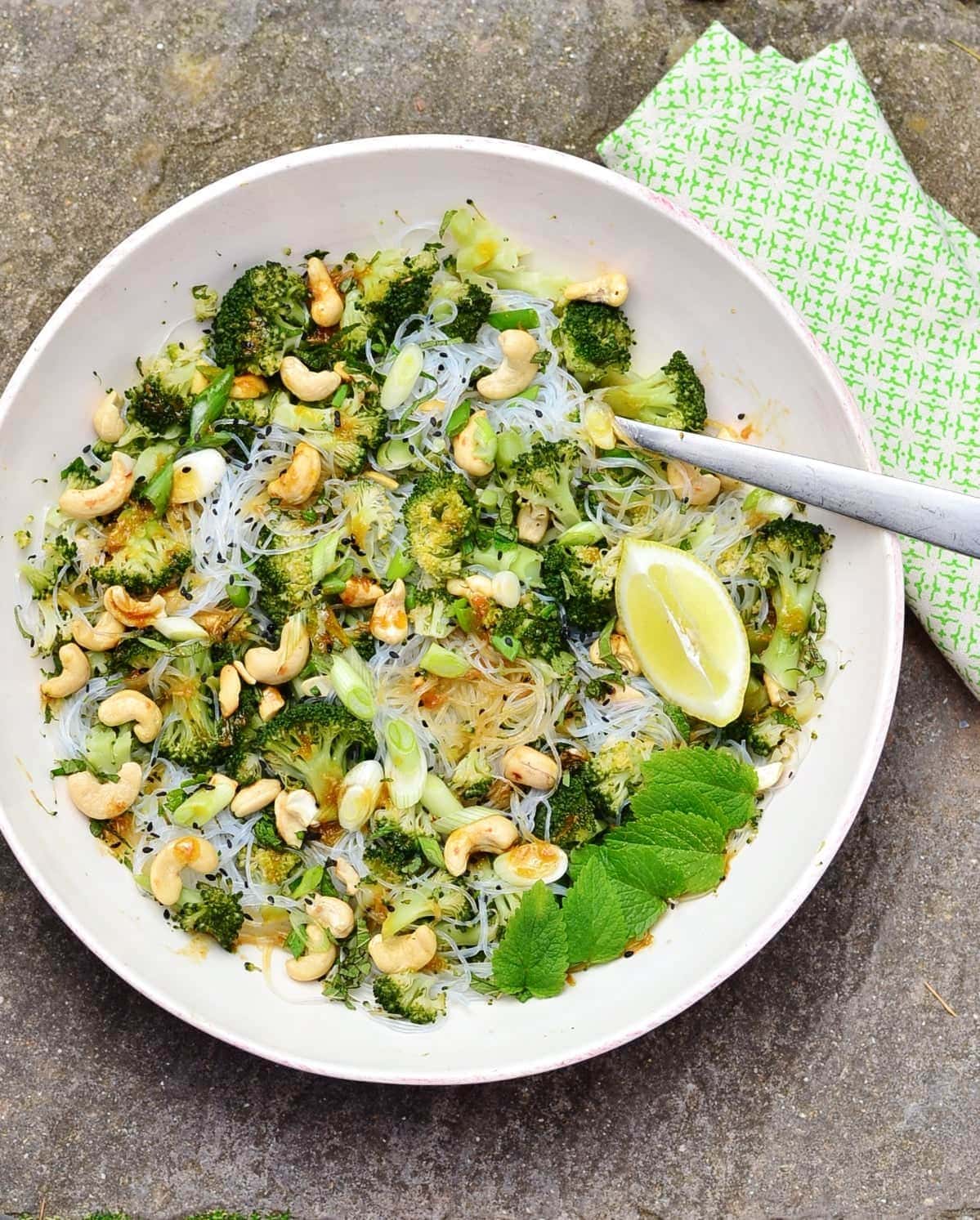 Top down view of bean thread noodles broccoli salad with cashews, lemon wedge, mint leaves and fork in white bowl with green cloth to right.