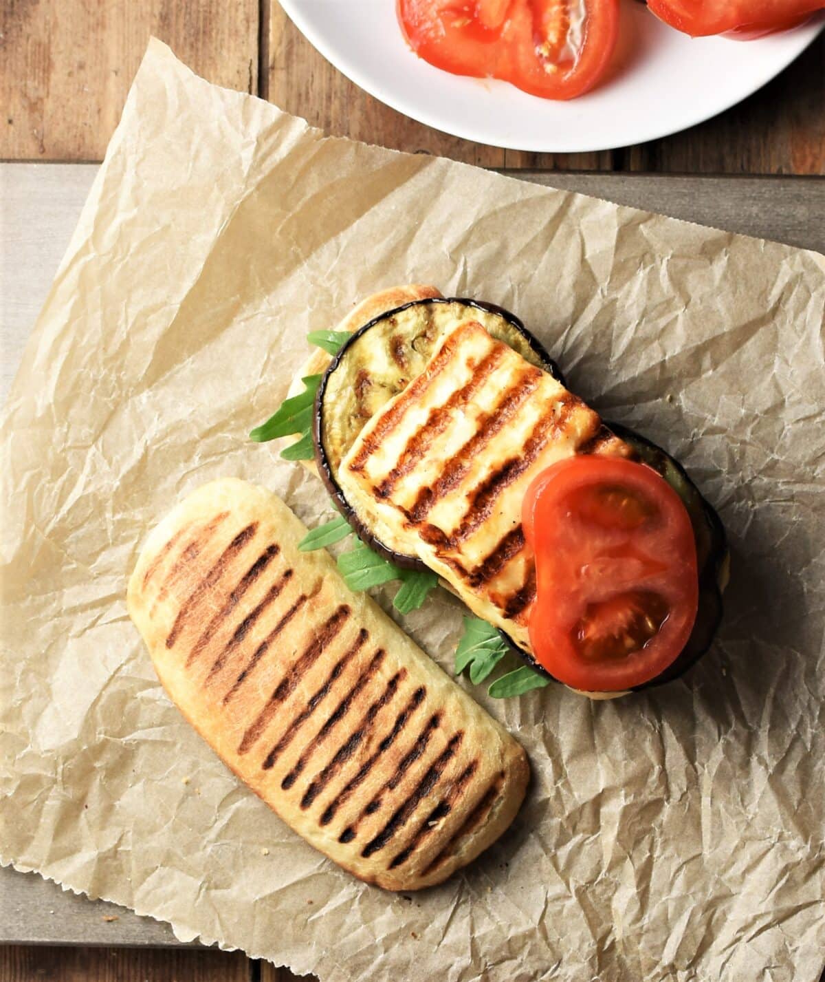 Top down view of open grilled eggplant and halloumi sandwich with tomato on top of paper.