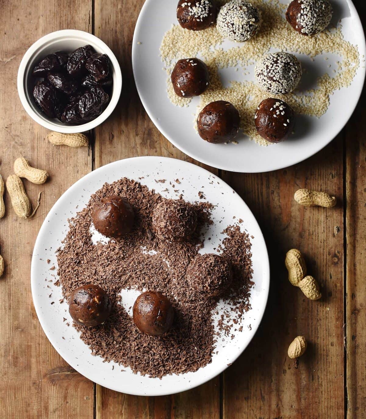 Energy balls on 2 white plates with seeds and chocolate flakes, with peanuts and prunes in small white container in background.