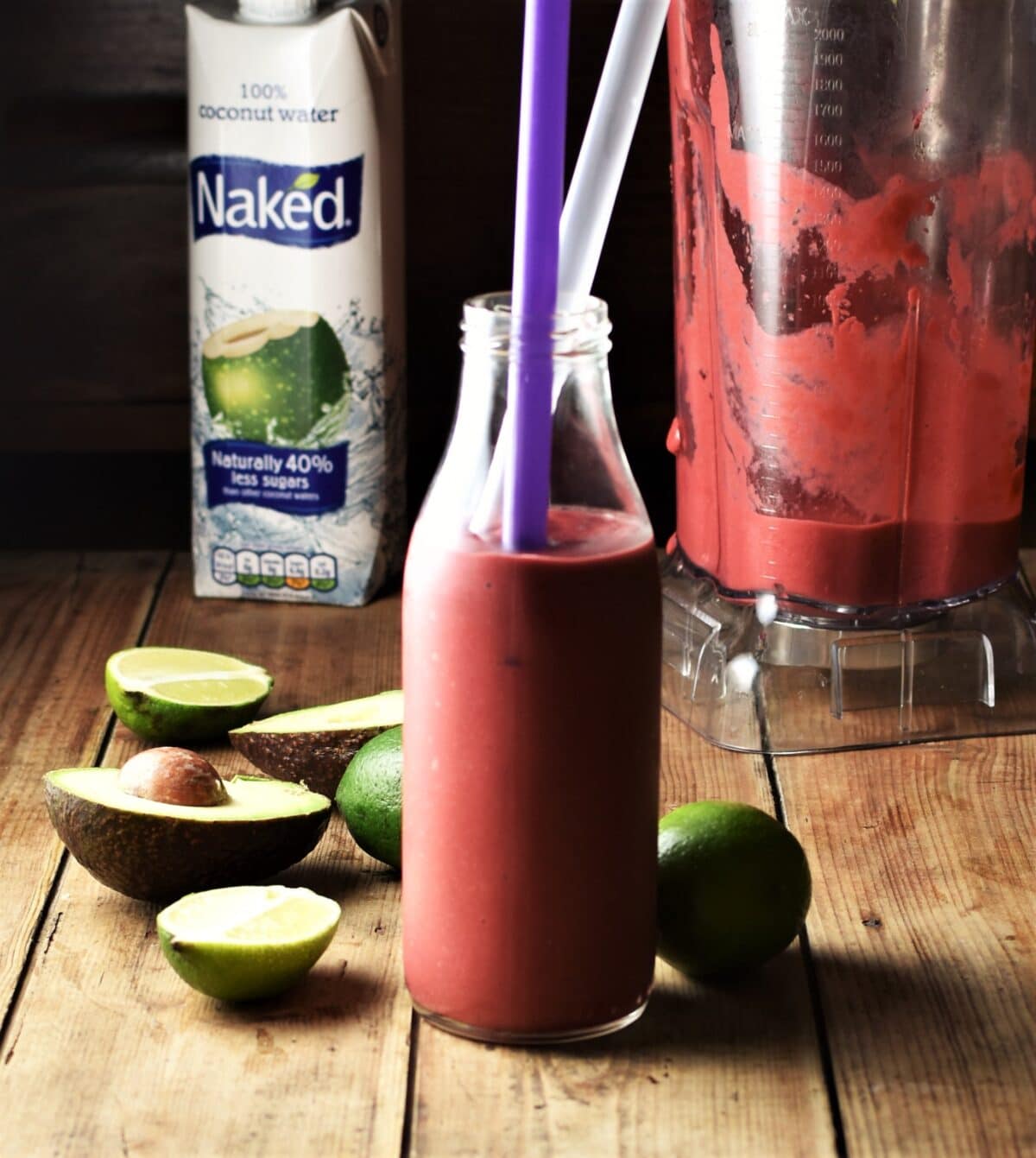 Side view of raspberry avocado smoothie in bottle with 2 straws, with limes, avocado, coconut water in carton and smoothie in blender in background.