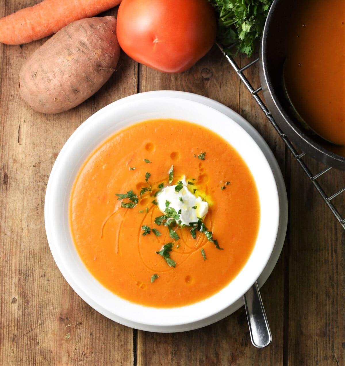 Creamy red vegetable soup in white bowl with sweet potato, carrot, tomato and pot in background.