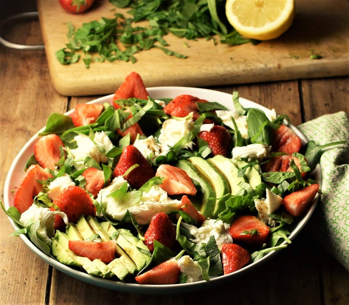 Avocado, spinach, strawberry and mozzarella salad on white plate with lemon and chopped spinach in background.