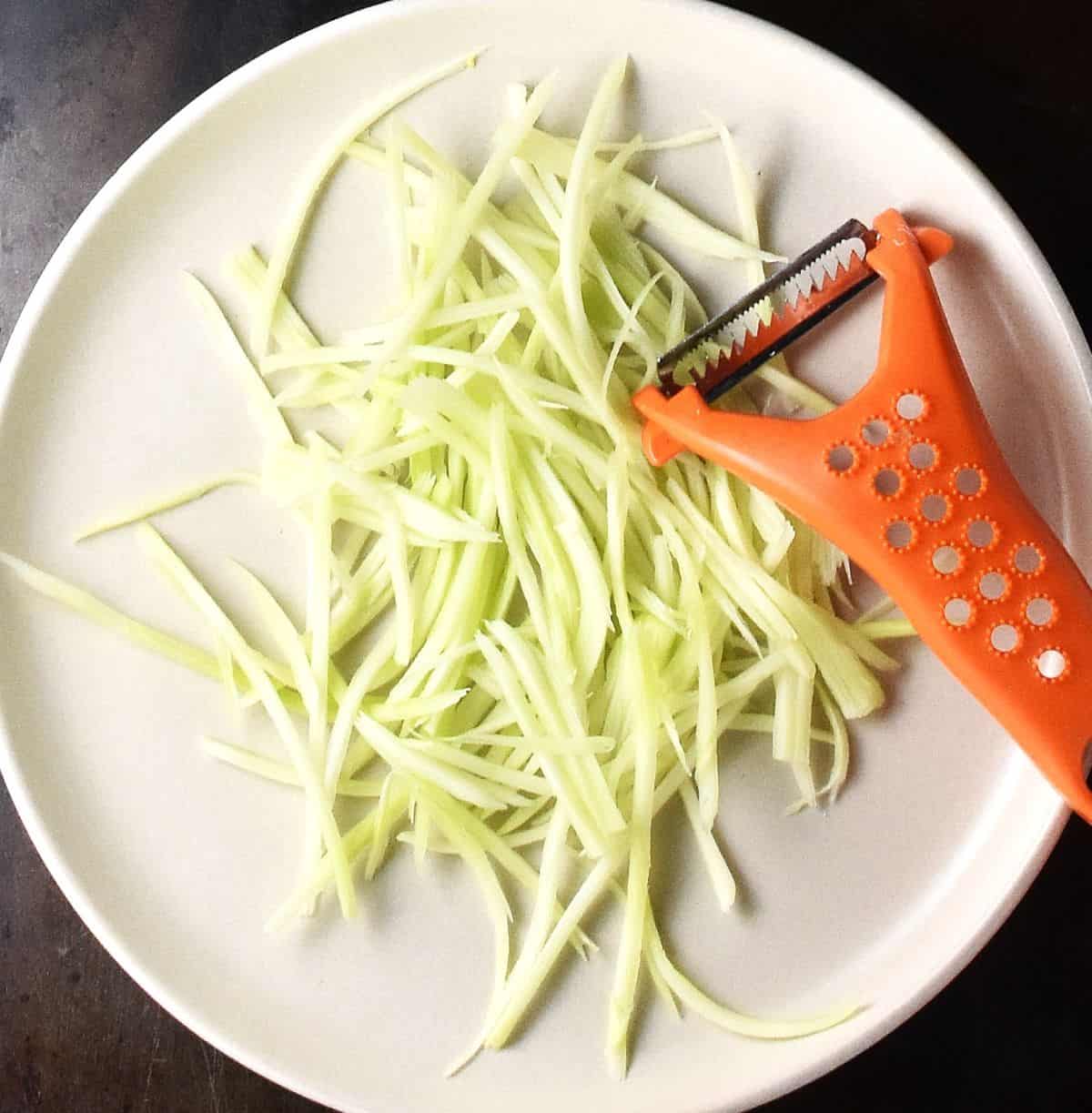 Julienned broccoli stalk on white plate with orange peeler.