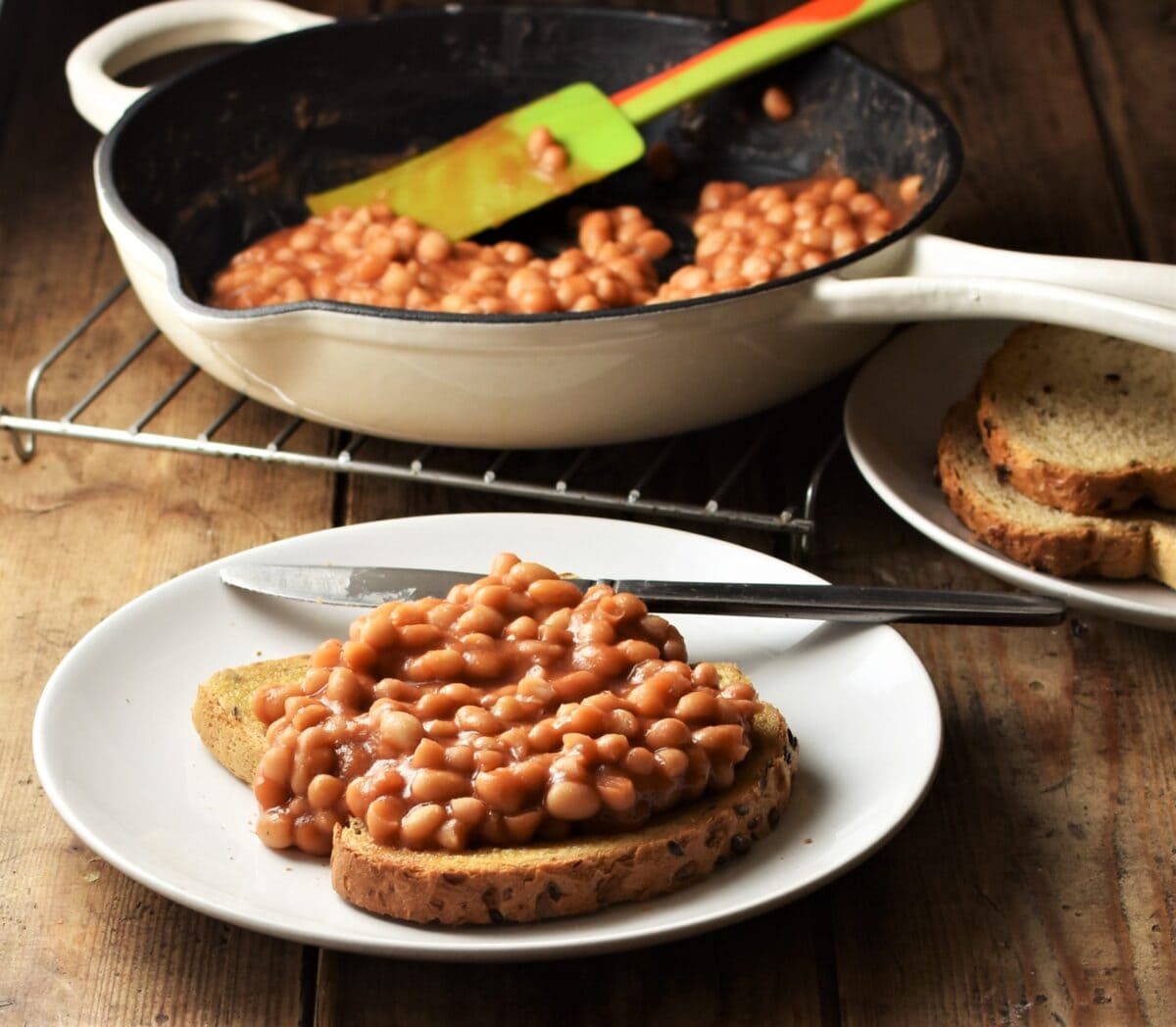 Side view of toast with baked beans and knife on white plate with beans in skillet in background.