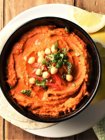 Top down view of red pepper hummus in black bowl on top of plate with tortillas and lemon wedges.
