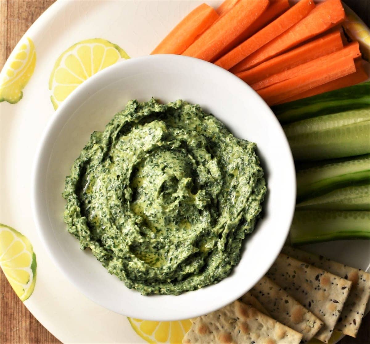 Creamy kale dip in white bowl with vegetable sticks and crackers in background.