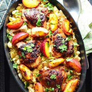 Top down view of crispy chicken with peaches on a bed of cauliflower and bulgur inside black oval casserole dish, with spoon on top of green cloth to the right.