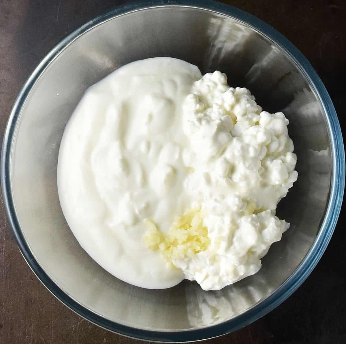 Top down view of cottage cheese, yogurt and chopped garlic in glass bowl.