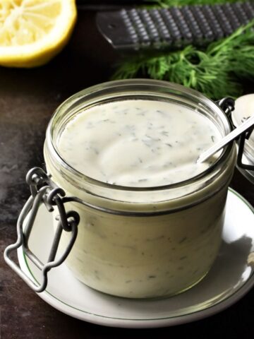 Healthy ranch dressing in open jar with spoon, lemon and herbs in background.