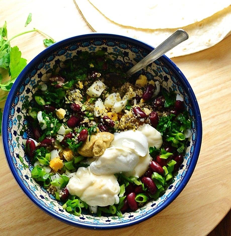 Top down view of mixture of egg, beans, onions, mustard, quinoa and yogurt in blue bowl with spoon on top of wooden surface with wraps at top