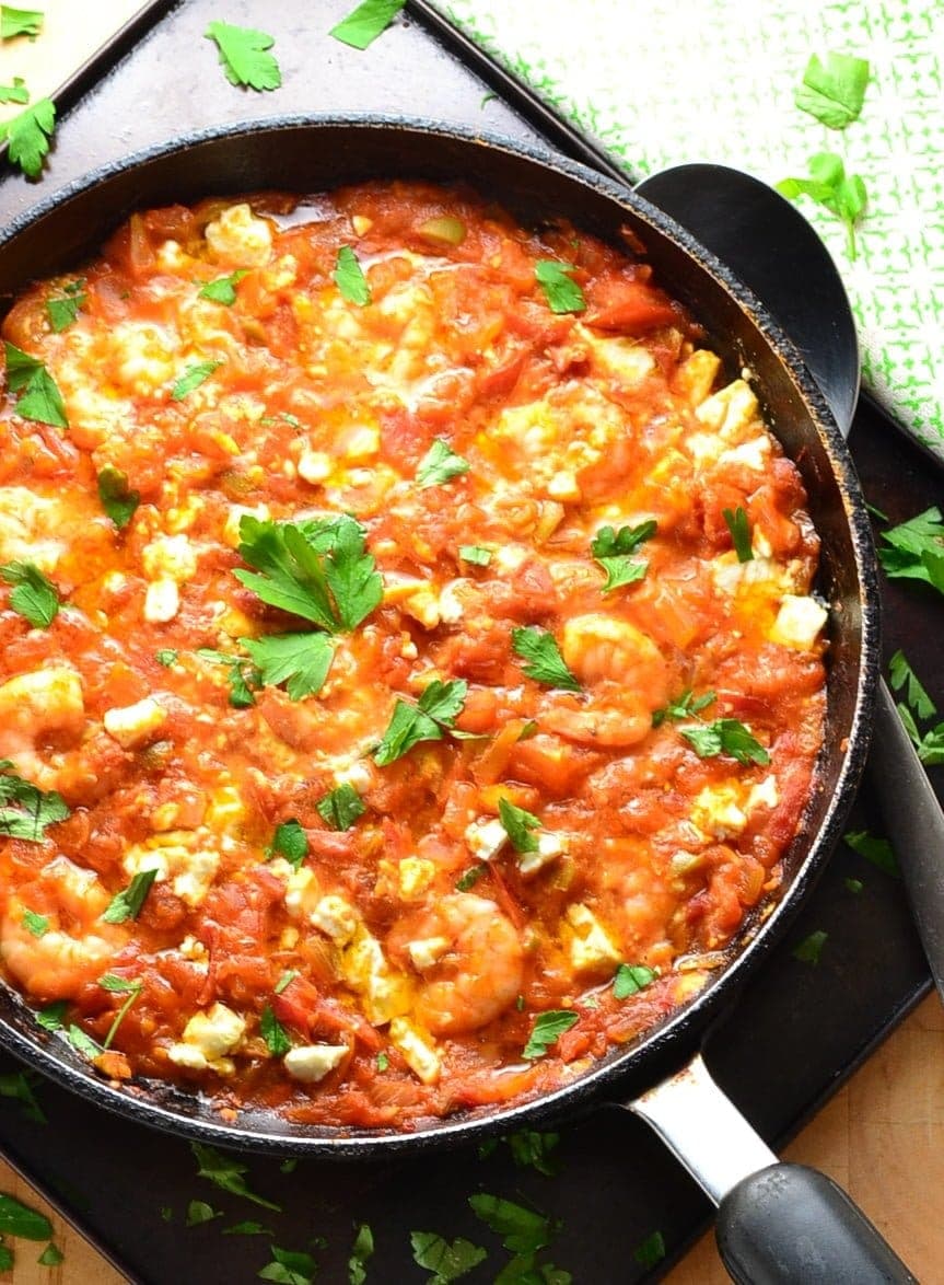 Top down partial view of shrimp tomato saganaki with herbs inside black skillet on top of dark tray, with large spoon and green cloth in top right corner.