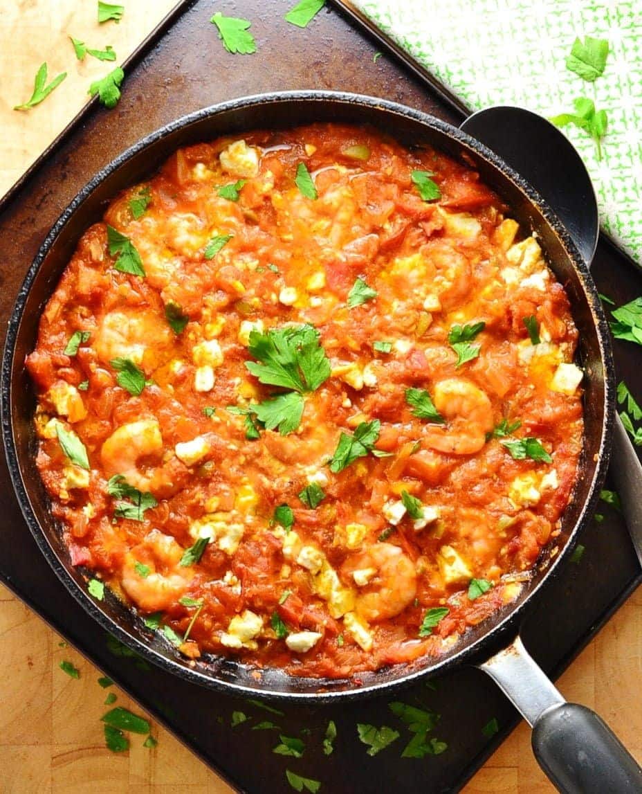 Greek shrimp saganaki in large black pan with handle and spoon on oven tray with green cloth.