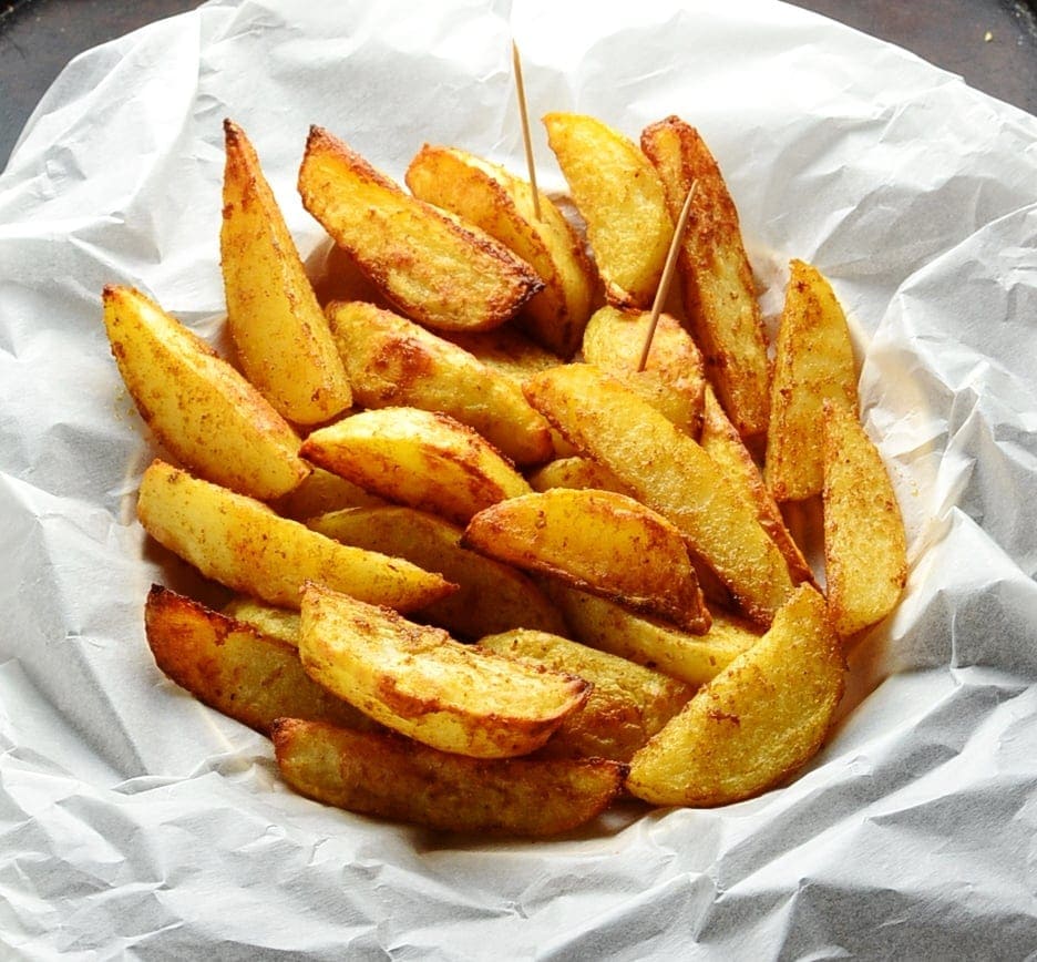 Spiced crispy potato wedges with toothpicks in basket lined with white paper.