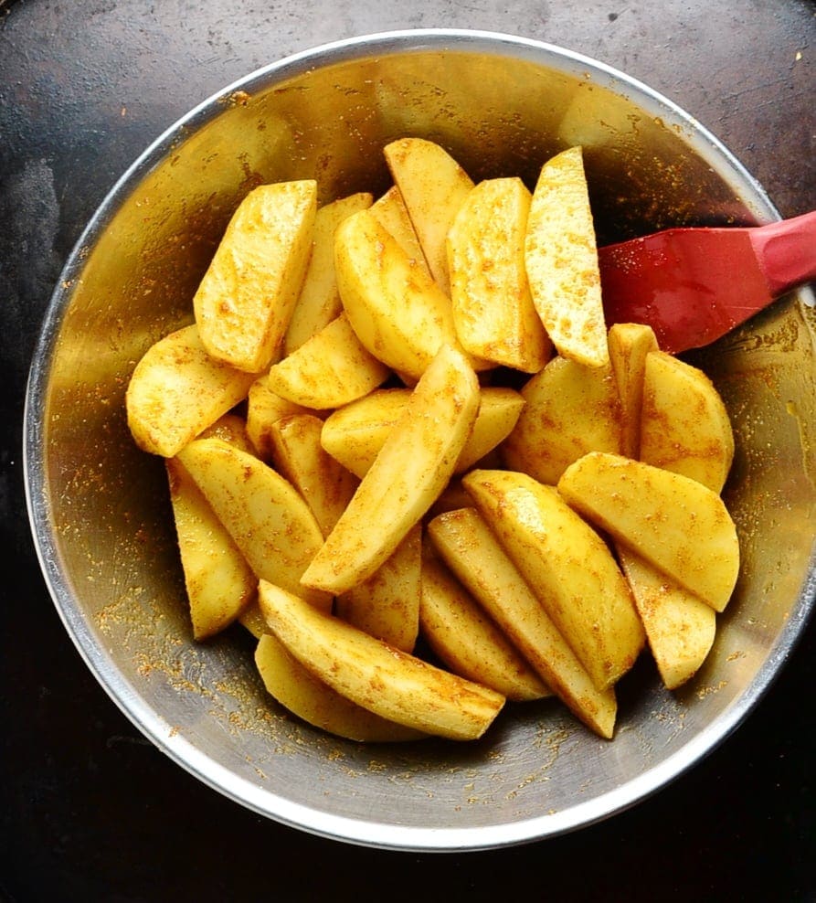 Raw spiced potato wedges in metal bowl with red spatula.