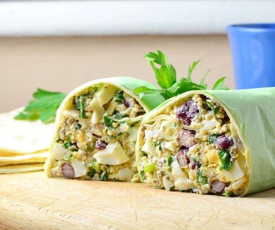 Close-up view of halved egg salad wrap on top of wooden board with parsley leaves and blue cup in background.