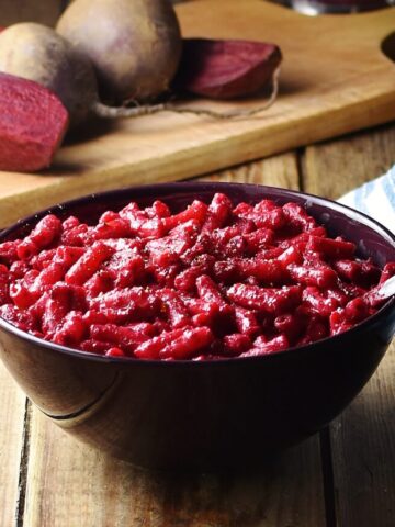 Beetroot pasta in purple bowl with spoon, cloth and raw beets in background.