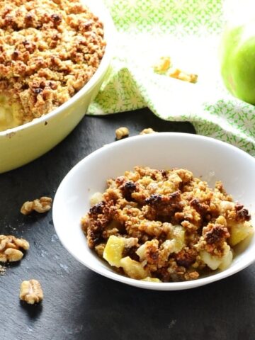 Apple crumble in white bowl, yellow dish with crumble and green cloth with green apple and spoon on top in background.