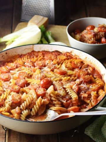 Fussili pasta with sliced fennel and tomato sauce in large white casserole dish with spoon, pasta and sauce in bowl, halved fennel and grater in background.