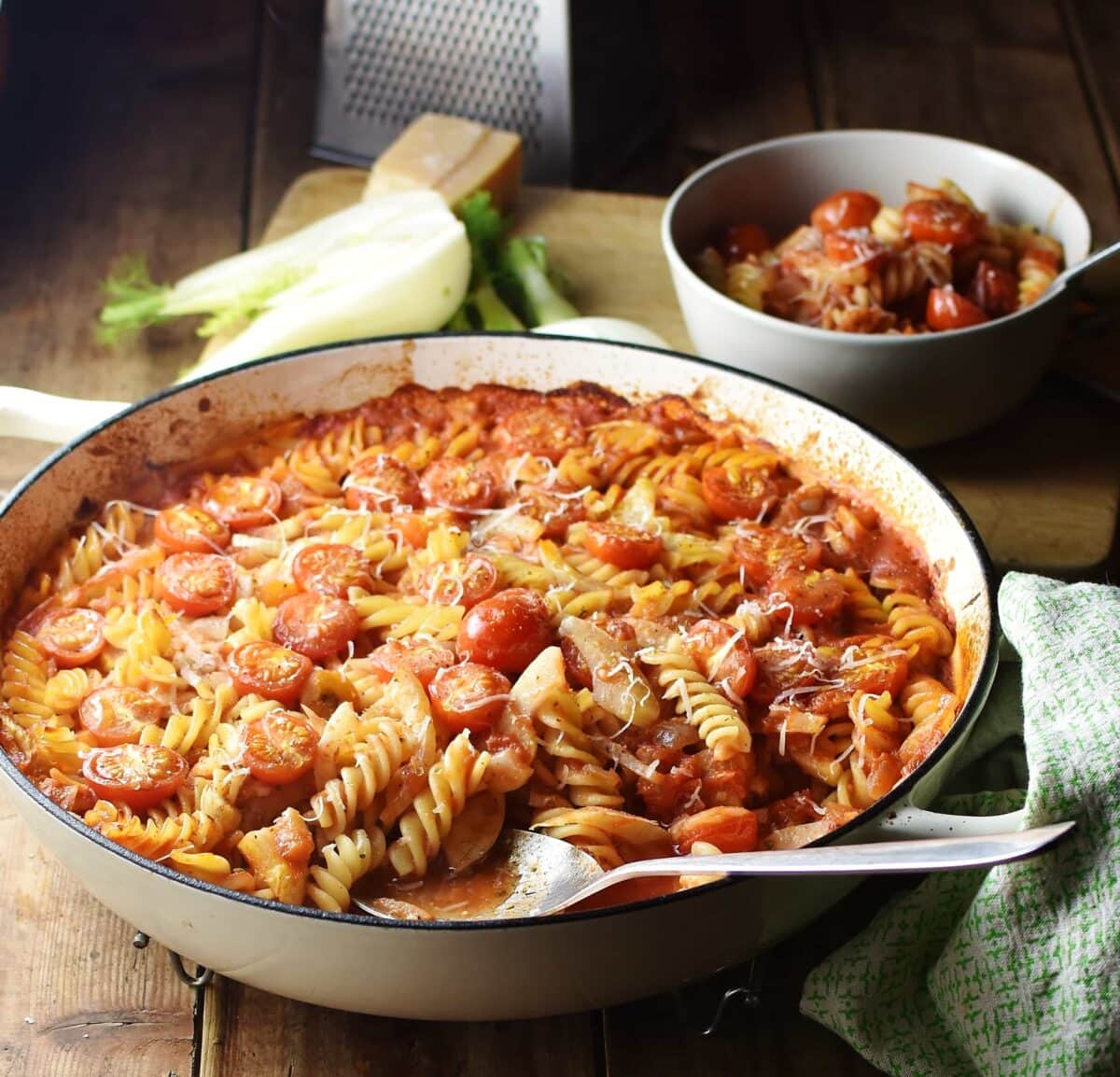 Fussili pasta with sliced fennel and tomato sauce in large white casserole dish with spoon, pasta and sauce in bowl, halved fennel and grater in background.