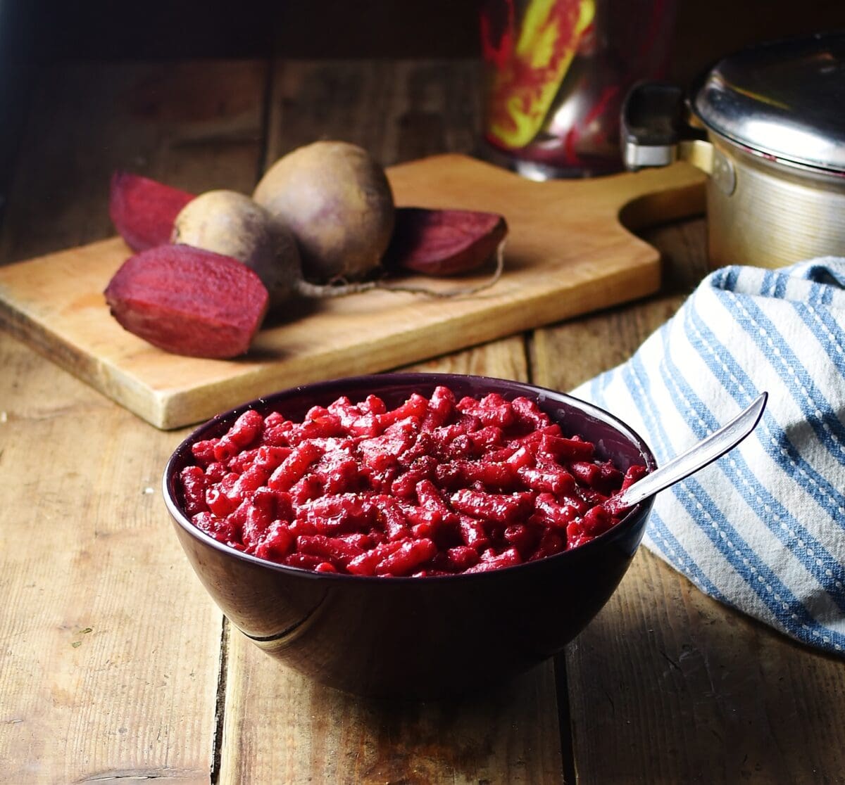 Beetroot macaroni in purple bowl with spoon, raw beets on wooden board and stripy blue cloth in background.