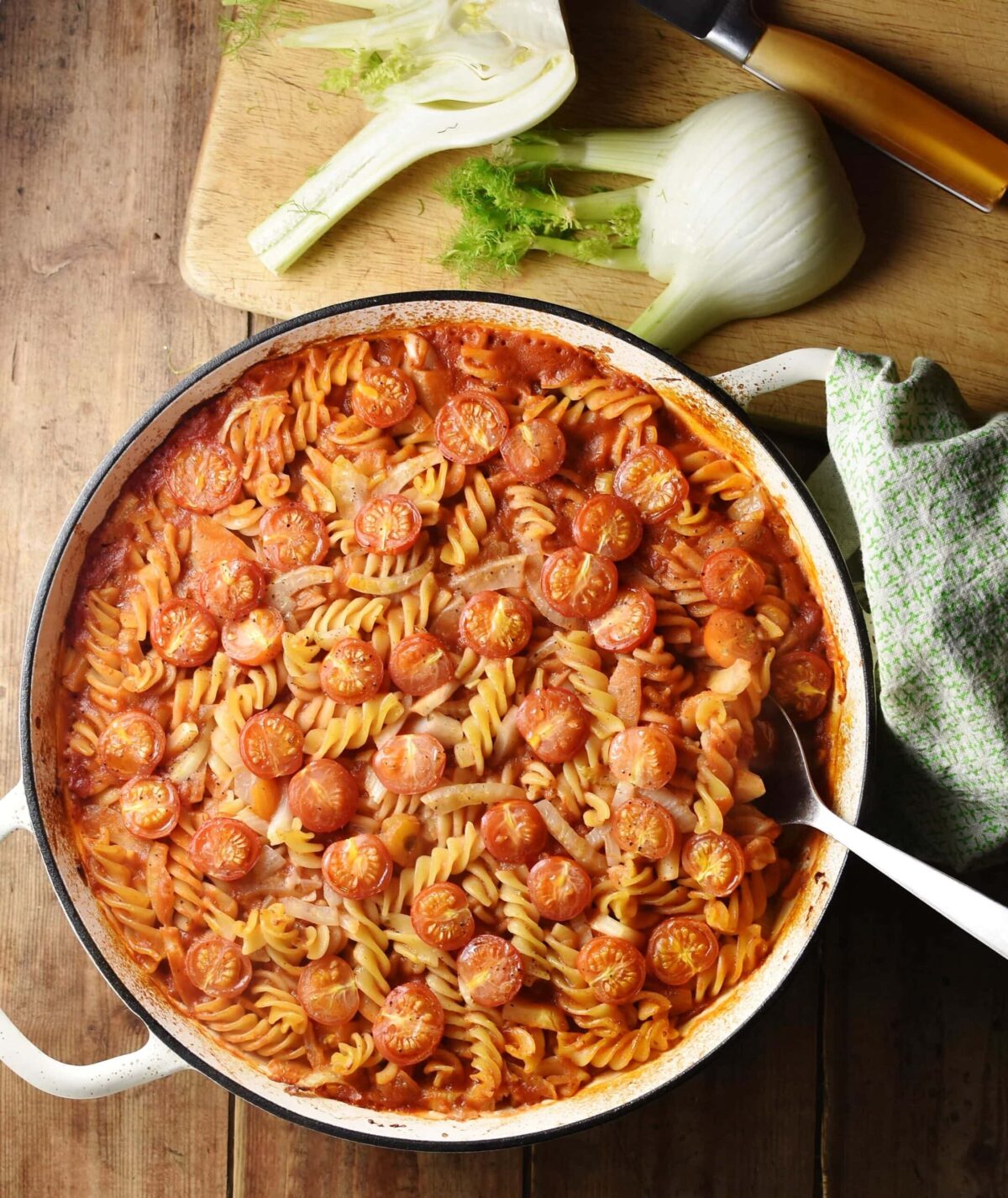 Pasta, fennel and tomatoes in large shallow, white dish with spoon, green cloth to the right and raw fennel with knife on top of wooden board in background.