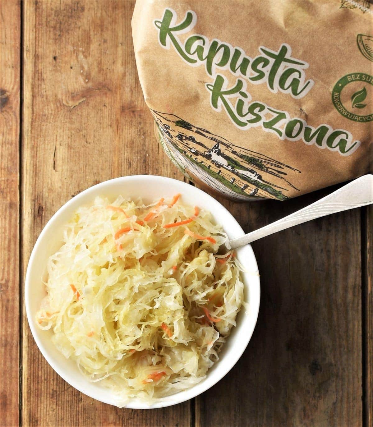 Top down view of sauerkraut in white bowl with spoon and sauerkraut packaging.