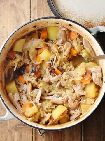 Potatoes, carrots, chicken pieces and sauerkraut in large white pot with spoon and lid in top right.