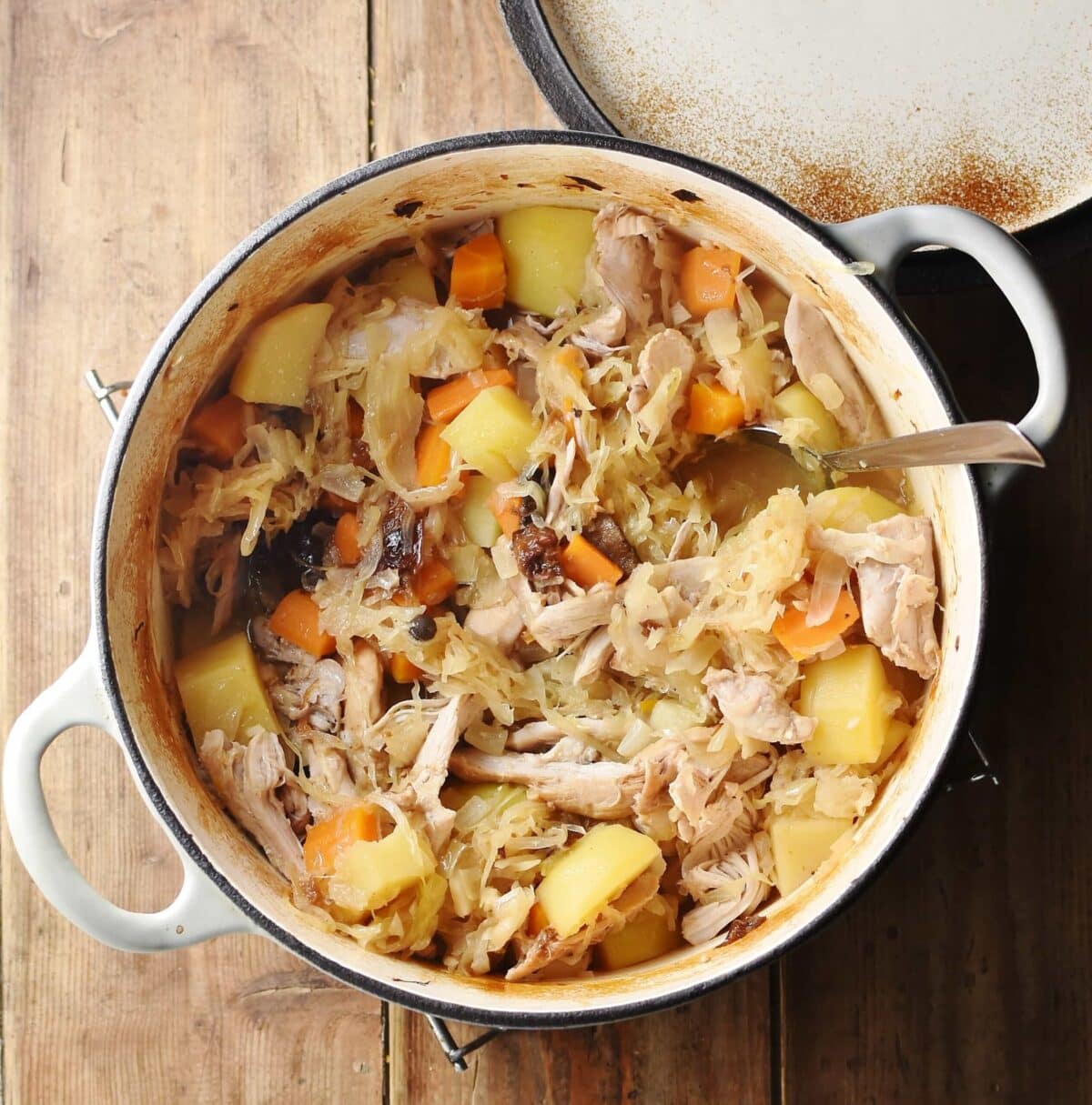 Potatoes, carrots, chicken pieces and sauerkraut in large white pot with spoon and lid in top right.