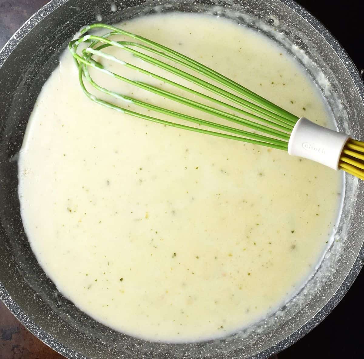 Creamy sauce in large, shallow pan with green whisk.