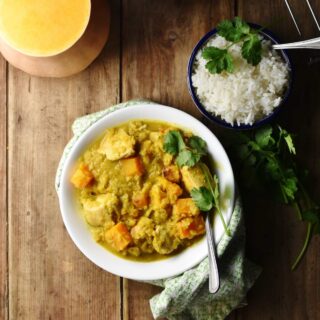 Chicken squash curry in white bowl with spoon wrapped in cloth, with rice in small bowl in top right and halved squash in top left.