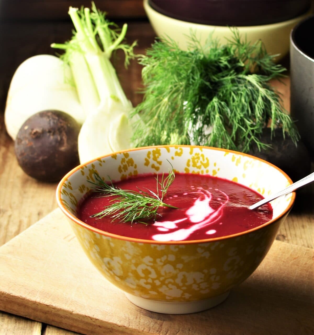 Side view of creamy beet soup with dill and yogurt in yellow patterned bowl with spoon, dill, fennel and bowls in background.