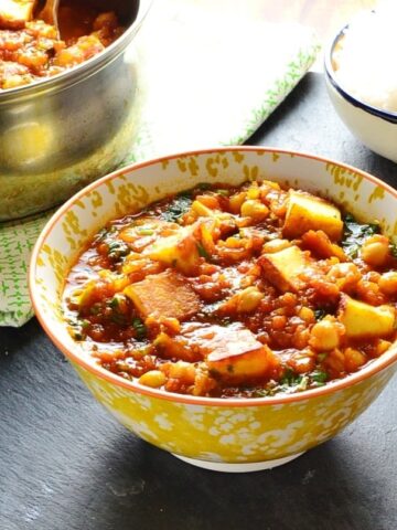 Vegetable curry with paneerin white-and-yellow bowl with saucepan and bowl of rice in background and fork to right.