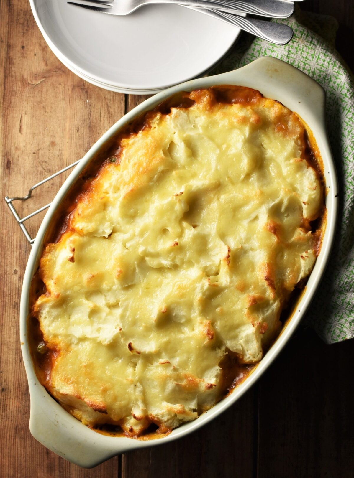 Pie with mashed potato topping baked golden in oval green dish, green cloth and white plates with forks in background.