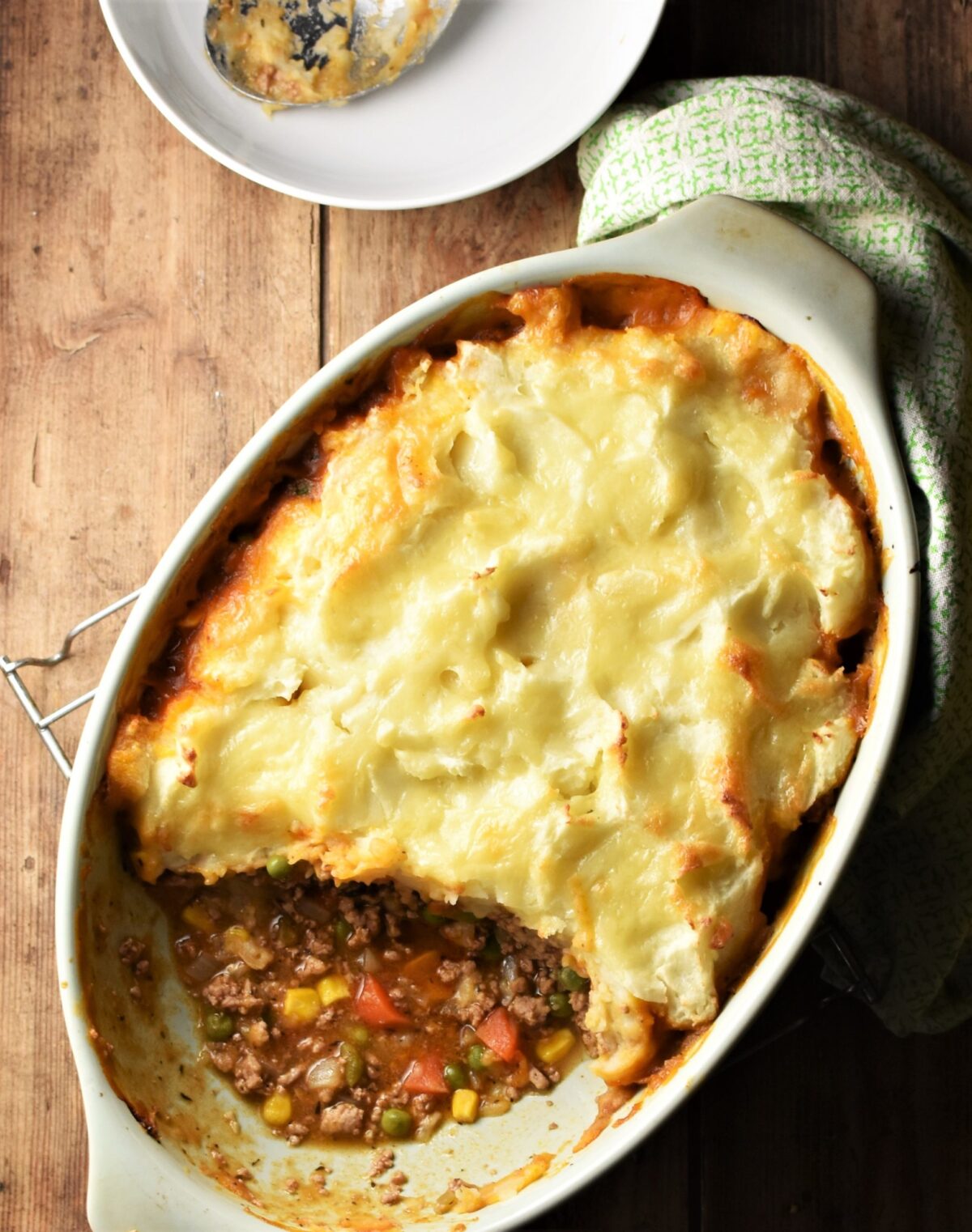 Top down view of turkey shepherds pie in green oval dish, with green cloth and white plate in background.