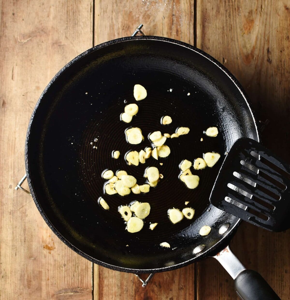 Slices of garlic with oil in pan with black spatula.
